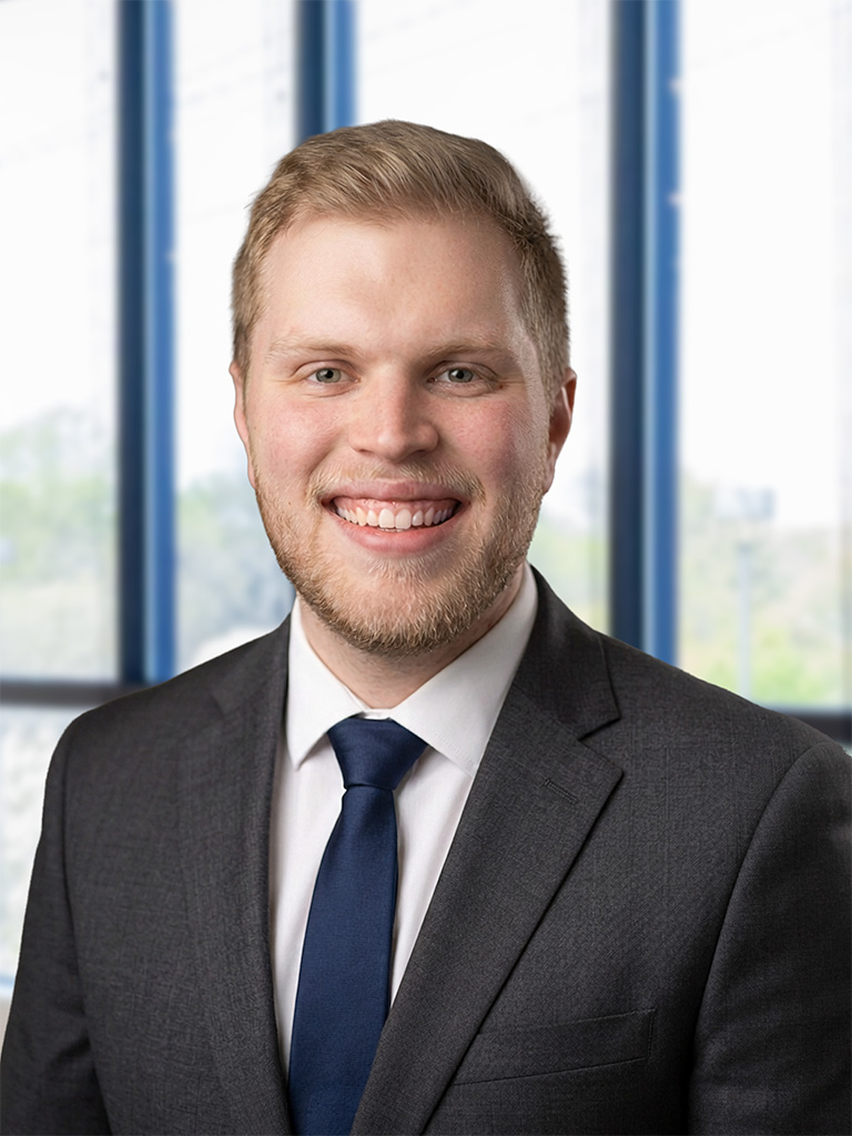 Professional headshot of a smiling man in a dark gray suit and blue tie, with an office backdrop in soft focus.